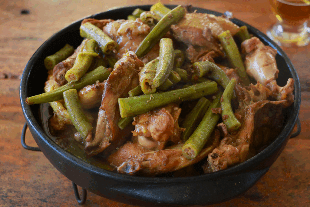 Frango caipira com quiabo servido em um tacho de ferro preto sobre mesa de madeira: pedaços de frango dourados e fatias de quiabo em um caldo encorpado, com uma dose de bebida ao fundo.