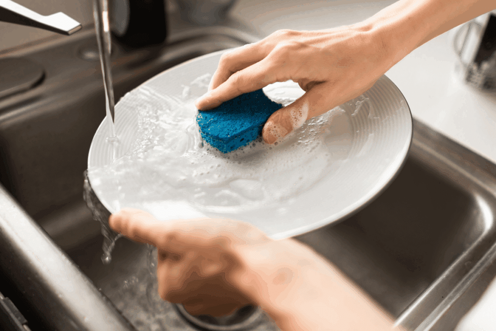 Mãos lavando um prato raso de cerâmica com esponja ensaboada sob água corrente na pia de inox