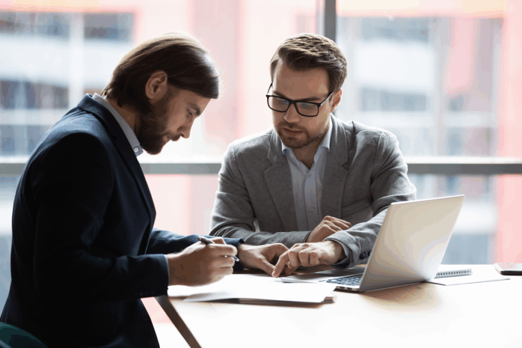 Dois homens empresários vestindo roupas formais em uma mesa, com notebook e papéis ao redor