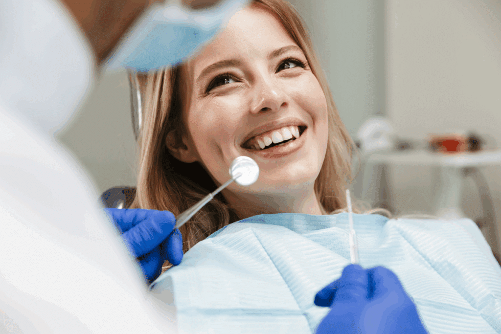 Uma jovem com cabelo loiro está sentada em uma cadeira odontológica, sorrindo enquanto recebe atendimento. Um profissional, parcialmente visível em primeiro plano, usa luvas azuis e examina a boca dela com um espelho odontológico. A paciente veste um avental descartável azul-claro, e o consultório aparece desfocado ao fundo.