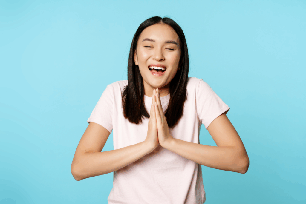 Uma mulher jovem asiática, com cabelos escuros e usando camiseta rosa clara, sorri alegremente de olhos fechados com as palmas das mãos unidas na altura do peito em gesto de agradecimento ou pedido. Ela está posicionada contra um fundo azul-claro vibrante.