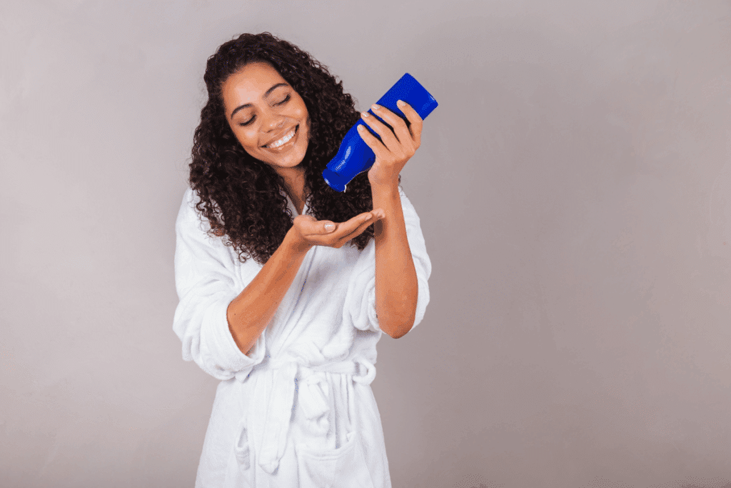 Mulher de cabelos cacheados escuros, sorrindo de roupão branco, colocando produto de um frasco azul na palma da mão para aplicar no cabelo.