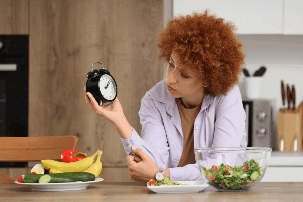 Fotografia mostra uma mulher de cabelos curtos, cacheados e ruivos, usando uma camisa lilás sobre uma blusa marrom, sentada à mesa em uma cozinha. Ela segura um relógio despertador preto com uma das mãos e observa o horário, enquanto uma tigela de salada, um prato com vegetais e uma seleção de frutas e legumes aparecem sobre a mesa.