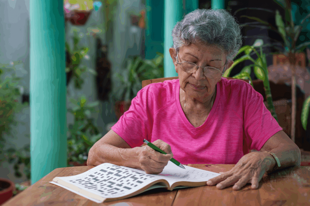 Mulher idosa de cabelos curtos, grisalhos e levemente ondulados, usando óculos de grau e camiseta rosa de manga curta. Ela está sentada à mesa e segura uma caneta verde enquanto escreve ou resolve uma atividade em uma folha de papel. A cena ocorre em um ambiente externo ou varanda, cercado por plantas e vasos, com colunas em tom verde-turquesa ao fundo. A mesa é de madeira e a composição tem iluminação natural.
