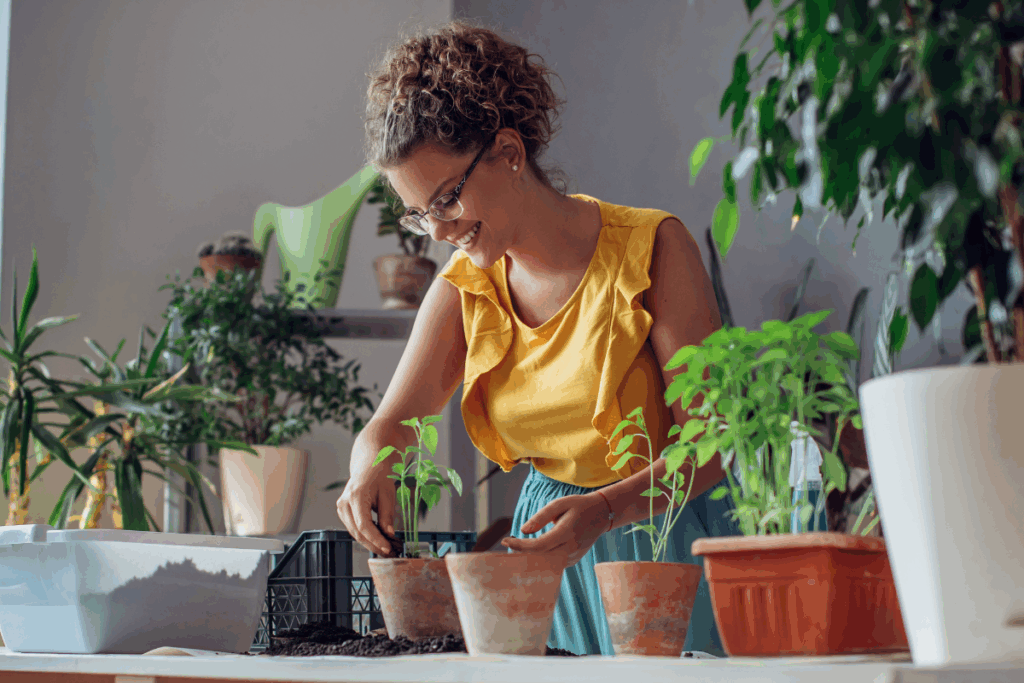 Fotografia de uma mulher adulta, com cabelos cacheados presos, usando óculos e uma blusa amarela sem mangas com babados. Ela aparece sorrindo enquanto cuida de pequenas plantas em vasos, em um ambiente interno repleto de vegetação. Sobre a mesa há vasos de diferentes tamanhos, terra, mudas e utensílios de jardinagem. A cena transmite uma sensação de contato com a natureza, tranquilidade e bem-estar, com iluminação natural e composição acolhedora.