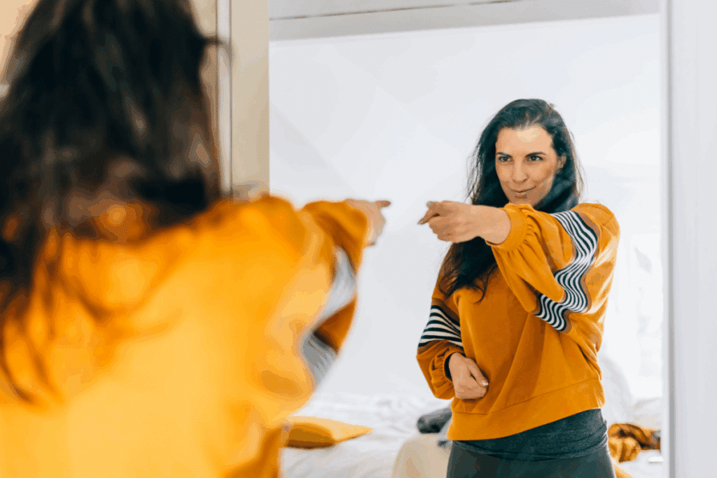 Mulher com cabelo liso, solto, usando blusa de moletom amarela com detalhes coloridos apontando para si mesma no espelho