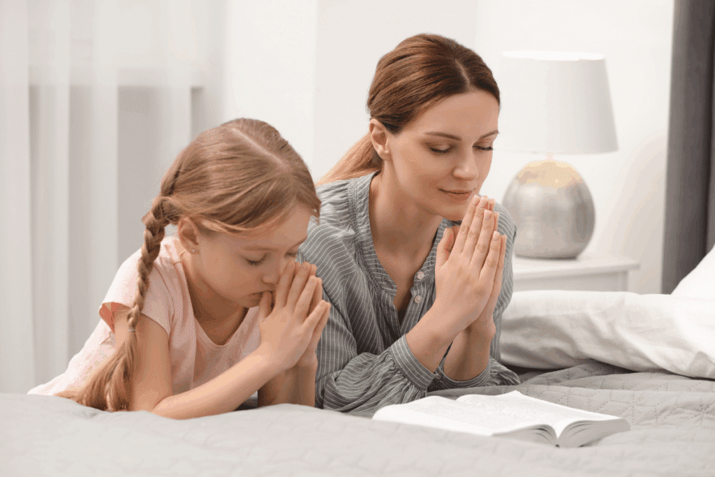 Uma mulher e uma criança deitadas em uma cama com as mãos postas em oração e os olhos fechados, tendo um livro aberto à frente delas