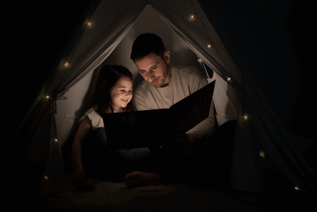 Pai e sua filha pequena sentados juntos dentro de uma pequena cabana ou barraca em formato de tenda, durante a noite. O pai tem cabelos castanhos-escuros, curtos, e barba curta. Ele veste uma camiseta de manga longa em tom bege-claro. A menina tem cabelos castanhos-escuros, longos e lisos, e veste uma blusa branca de manga curta com detalhes nas mangas. Os dois estão próximos, olhando para um livro ilustrado aberto que seguram juntos. A criança demonstra interesse e sorri enquanto acompanha a leitura, enquanto o pai olha atentamente para o livro. A tenda é escura por dentro e decorada com pequenas luzes amareladas, criando uma atmosfera aconchegante e intimista. A iluminação é baixa e quente, concentrada principalmente nos rostos e no livro.