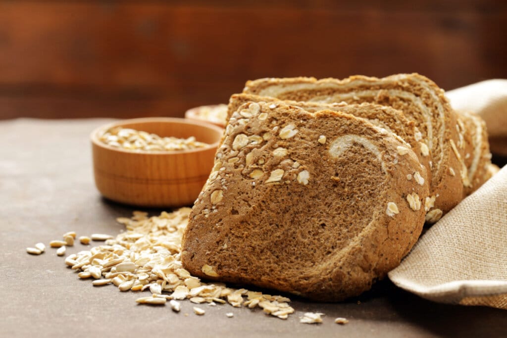 Fatias de pão integral de forma com grãos de aveia salpicados na casca, apoiadas lado a lado sobre uma mesa rústica. Ao fundo, pequenos recipientes de madeira e grãos soltos espalhados completam a composição.