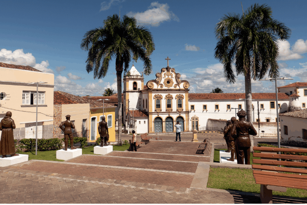 Praça no centro histórico de Penedo (AL), com estátuas de bronze no primeiro plano e a Igreja e Convento de Santa Maria dos Anjos ao fundo, sob céu ensolarado