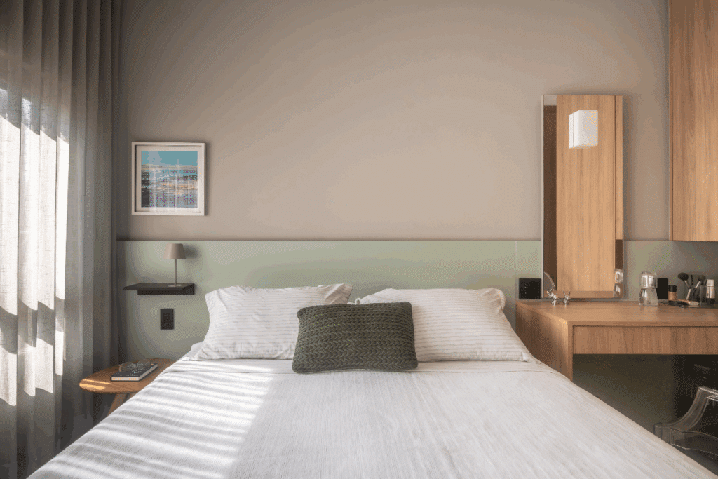 Um quarto moderno e iluminado, com uma cama de casal arrumada com roupas de cama brancas e uma almofada verde ao centro. A decoração combina tons neutros, verde-claro e madeira. À direita, há uma bancada com espelho e objetos pessoais; à esquerda, uma janela com cortinas permite a entrada de luz natural.