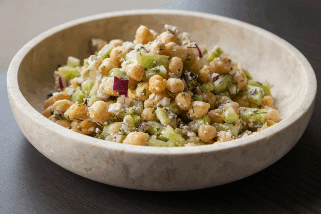 Salada de grão-de-bico com queijo feta, pepino e cebola-roxa servida em tigela de cerâmica em cima de mesa de madeira