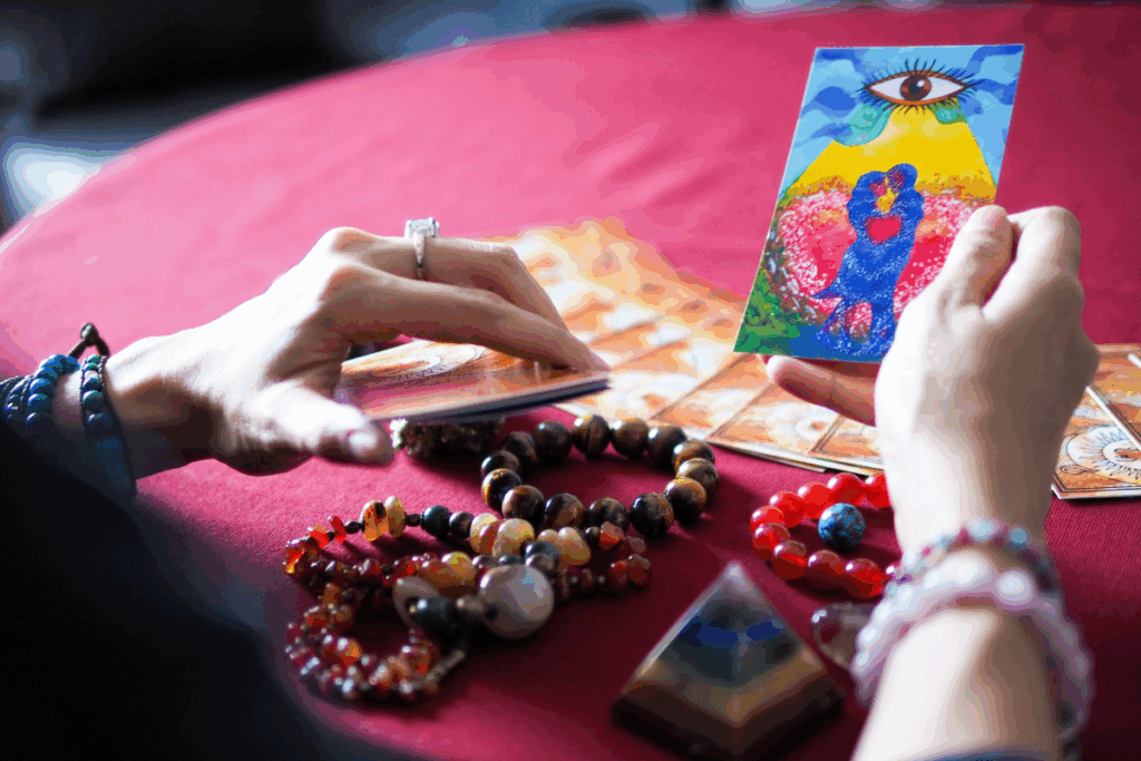 Visão em primeira pessoa das mãos de uma taróloga segurando cartas sobre uma mesa com toalha vermelha. Na mão direita, ela exibe uma carta ilustrada com um casal abraçado sob um grande olho e um céu azul; na mesa, há várias pulseiras de pedras coloridas, uma pequena pirâmide de cristal e outras cartas espalhadas