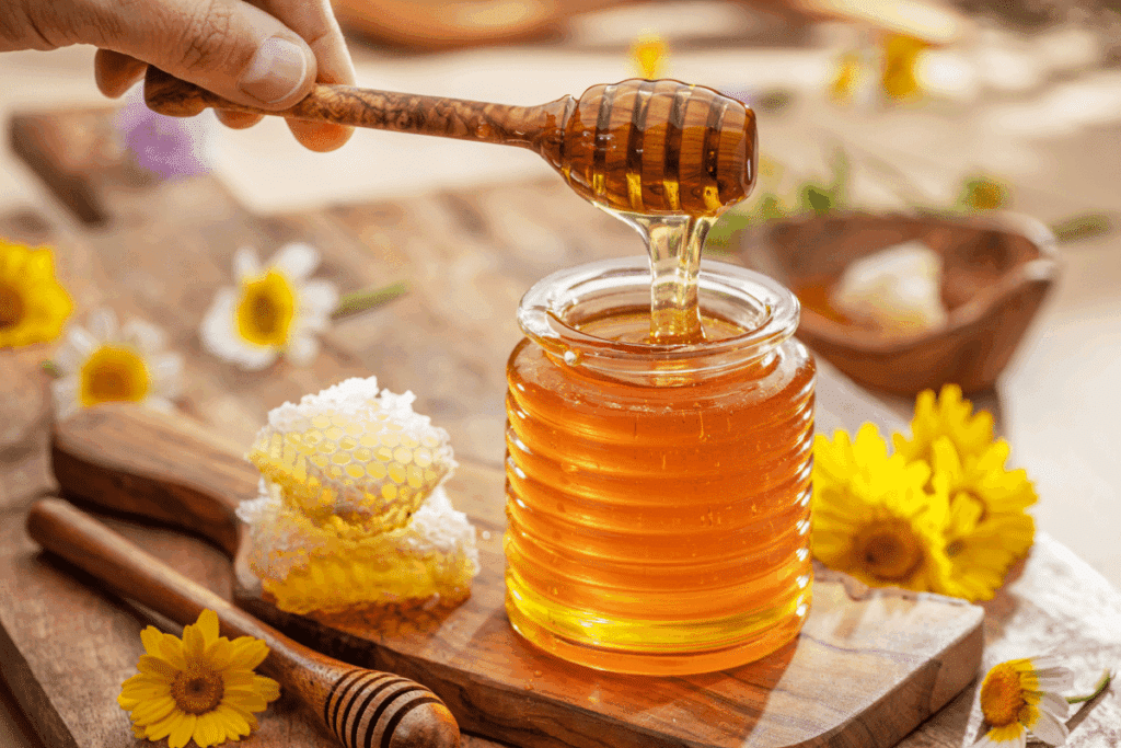 Mão segurando uma colher de mel sobre um pote de vidro cheio de mel dourado, ao lado de um pedaço de favo e flores amarelas e brancas sobre uma tábua de madeira.