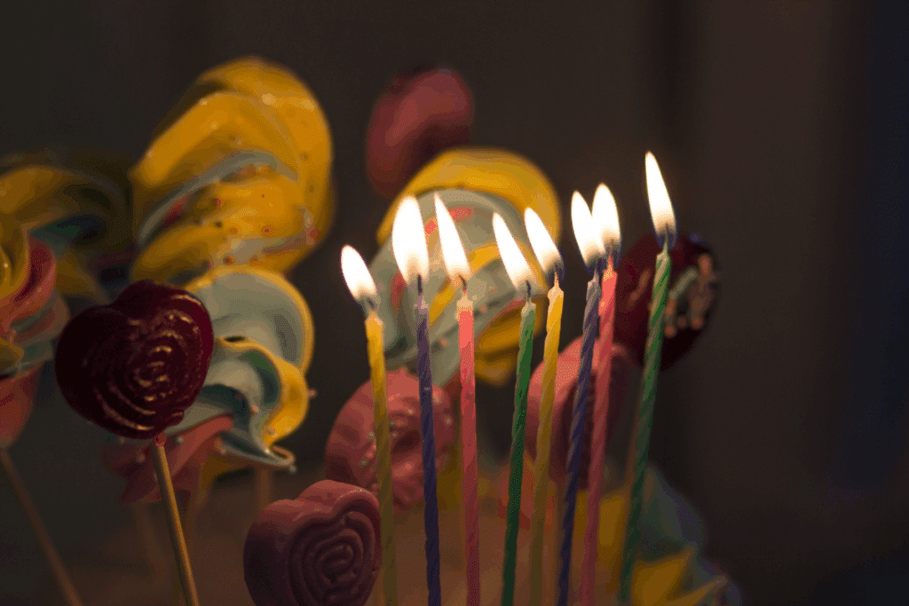Várias velas coloridas de aniversário acesas em primeiro plano, acompanhadas ao fundo por pirulitos e doces decorativos em tons pastéis