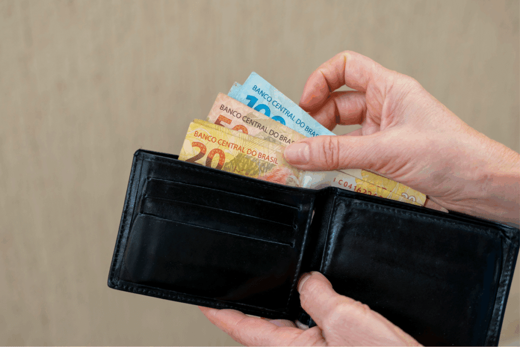 Mãos segurando uma carteira de couro preta aberta, exibindo cédulas de Real brasileiro