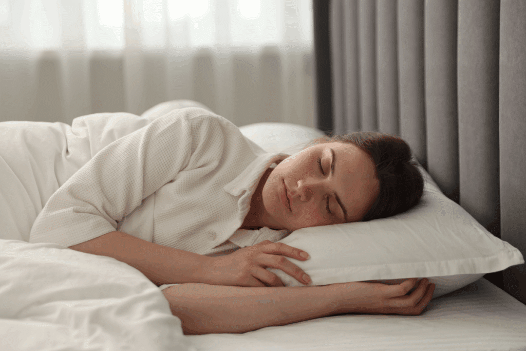 Mulher dormindo tranquilamente de lado na cama, com a cabeça apoiada em um travesseiro branco e roupa de cama clara, em um quarto com cortinas cinza.