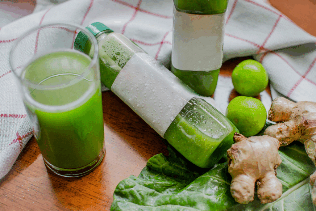 Um copo e duas garrafas plásticas transparentes com rótulos brancos exibem suco verde de couve sobre uma superfície de madeira. Ao lado dos recipientes, aparecem os ingredientes frescos: uma folha grande de couve, dois limões inteiros e pedaços de gengibre, sob um pano de prato quadriculado ao fundo