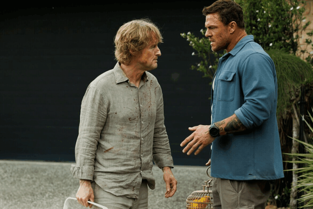 Cena do filme "Três Horas Para Viver": à esquerda, o ator Owen Wilson, de cabelos loiros e camisa social bege manchada de sangue, encara com expressão preocupada o ator Alan Ritchson, à direita, que veste uma camisa azul e tem uma tatuagem no antebraço. Os dois estão ao ar livre, diante de uma parede escura e plantas.