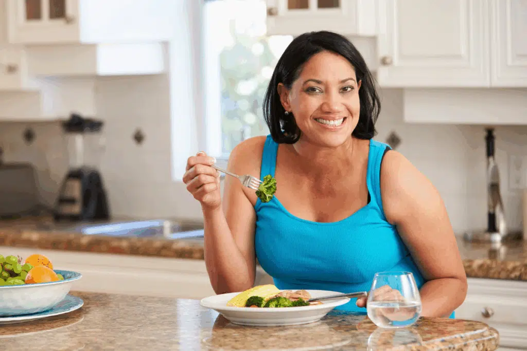 Mulher sentada à mesa em uma cozinha, sorrindo enquanto segura um garfo com um pedaço de brócolis. Ela tem o cabelo curto preto e veste uma regata azul e tem um prato com vegetais à sua frente, além de uma tigela com frutas ao lado.