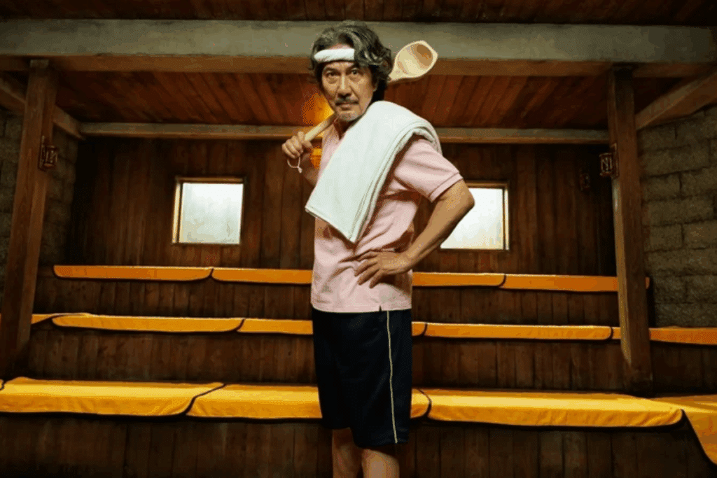 Um homem de cabelos grisalhos e faixa branca na cabeça segura uma grande colher de madeira no ombro e uma toalha sobre a outra parte da jaqueta, posando em uma sauna de madeira.