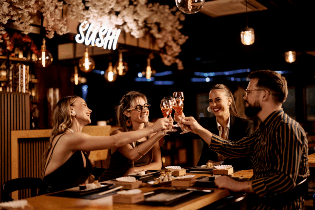 Fotografia de quatro amigos reunidos ao redor de uma mesa em um restaurante japonês, celebrando com um brinde de taças. A mesa está servida com pratos de sushi e sushis variados. O ambiente possui iluminação quente e acolhedora, com lâmpadas pendentes de filamento e um letreiro luminoso em neon com a palavra "SUSHI" ao fundo.