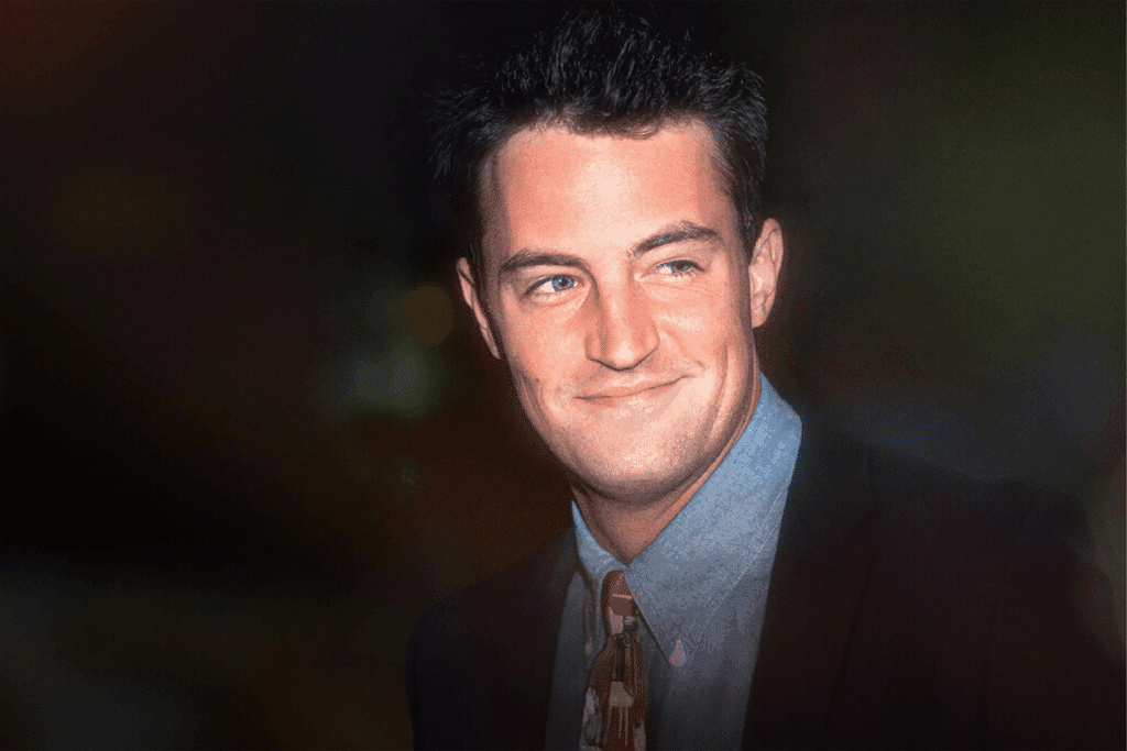Retrato de Matthew Perry sorrindo, vestindo camisa azul e terno escuro.