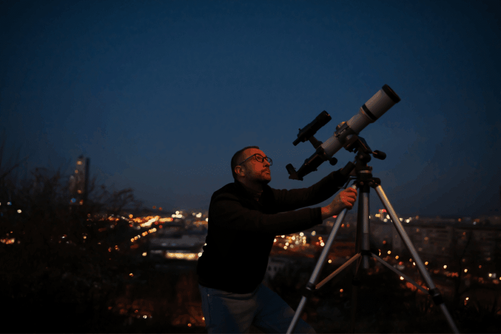 Um homem de óculos e casaco escuro ajusta um telescópio montado em um tripé durante a noite, com o céu escuro ao fundo e as luzes desfocadas de uma cidade ao longe.   