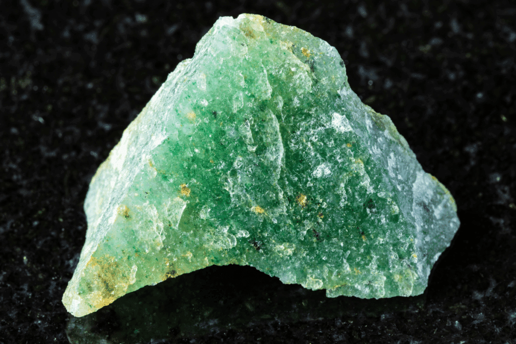 Pedra bruta de aventurina verde com formato triangular e textura irregular, apoiada sobre uma superfície preta.