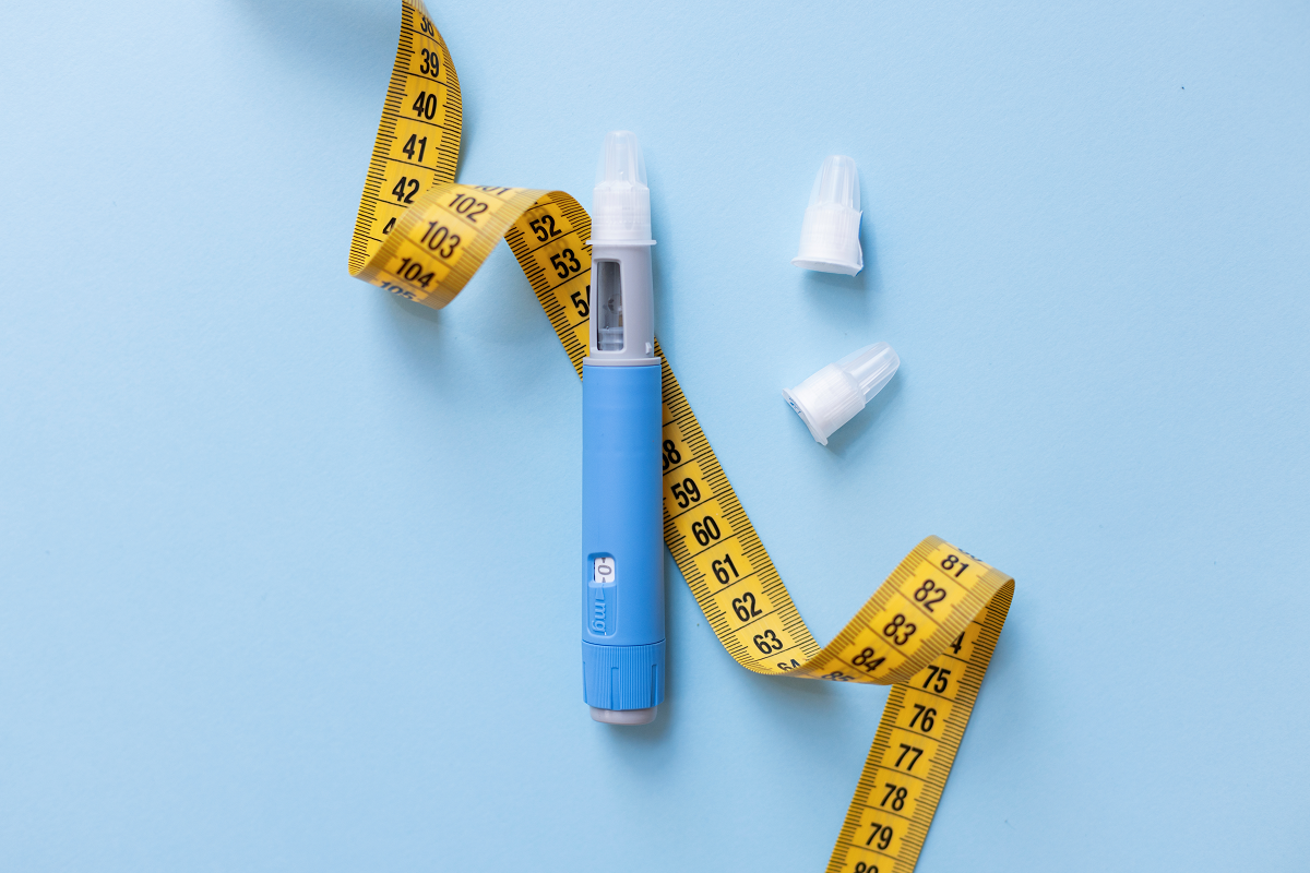 Uma caneta aplicadora de medicamento azul e cinza posicionada verticalmente no centro de um fundo azul-claro. Uma fita métrica amarela ondulada contorna a caneta, e duas tampas protetoras plásticas estão ao lado direito.