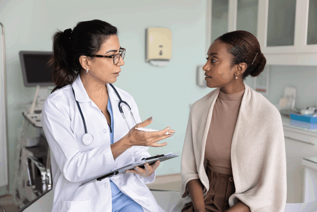 Médica de jaleco branco, óculos e estetoscópio gesticula enquanto conversa com uma paciente negra sentada à sua frente, em um consultório médico bem iluminado.
