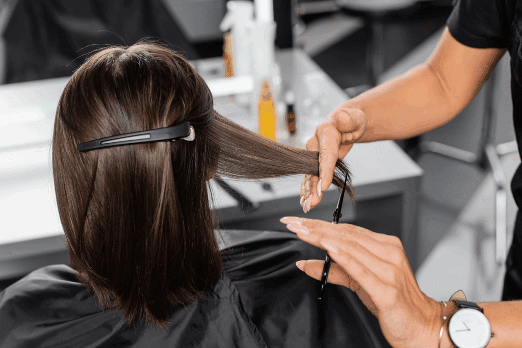 Plano detalhado mostrando as mãos de uma profissional cortando as pontas de uma mecha de cabelo castanho escuro com tesoura, enquanto o restante dos fios está preso por uma presilha preta.