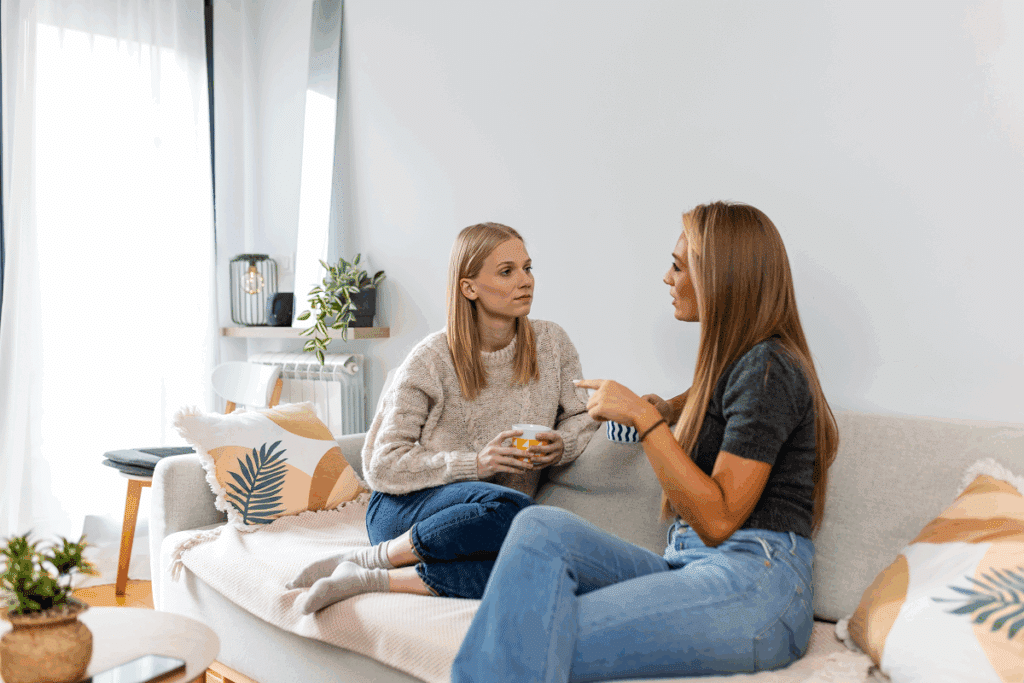 Duas mulheres sentadas em um sofá claro conversando e segurando canecas em uma sala iluminada e bem decorada.   