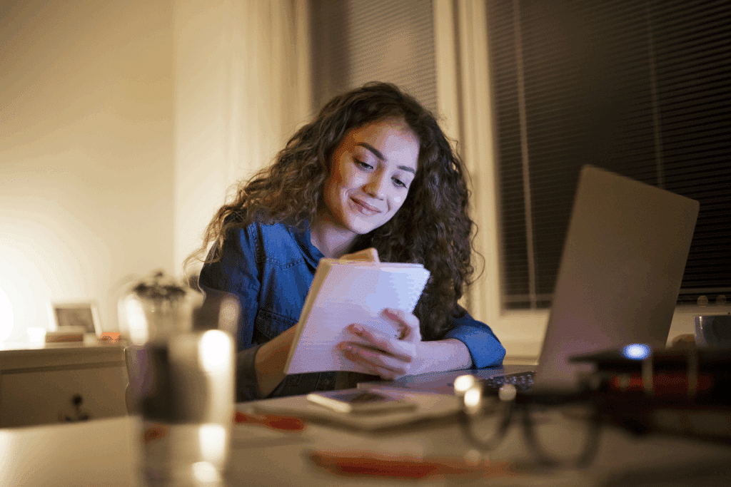 Jovem branca de cabelos cacheados sorrindo enquanto escreve em um bloco de anotações em frente a um notebook aberto, em um ambiente ilimitado à noite.