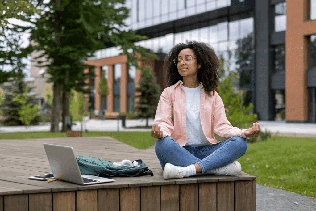 Jovem negra de óculos e camisa rosa sentada em posição de meditação de pernas cruzadas sobre um deck de madeira ao ar livre, com um notebook aberto e uma mochila verde ao seu lado.