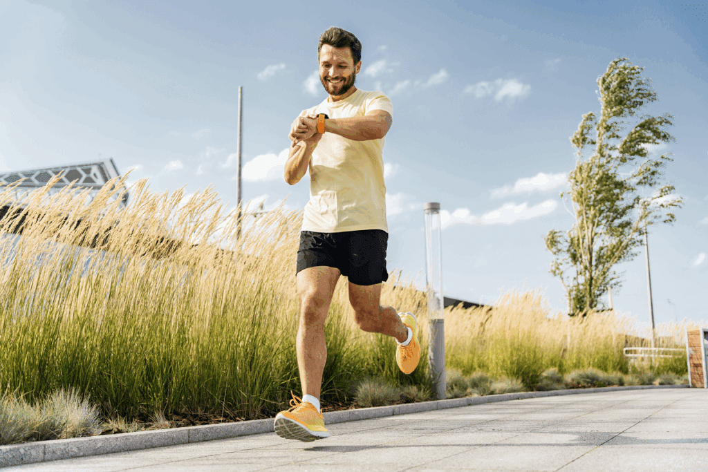 Um homem sorridente corre ao ar livre em uma calçada ensolarada olhando para o relógio em seu pulso. Ele veste camiseta amarela clara, shorts preto, tênis amarelos e pulseira laranja no relógio. Ao fundo, há grama alta, uma árvore e o céu ensolarado.   
