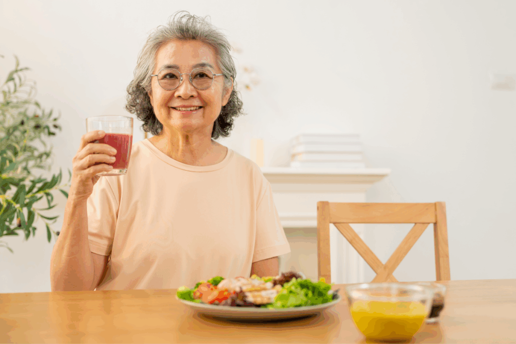 Uma mulher idosa de cabelos grisalhos e óculos sorri para a câmera enquanto segura um copo com suco vermelho. Ela está sentada à mesa de jantar, que contém um prato de salada colorida com frango e uma tigela de molho. Ao fundo, o ambiente é claro e bem iluminado.