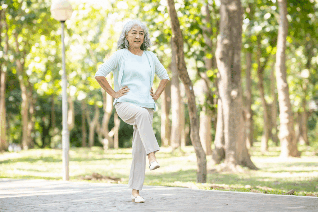 Mulher idosa com cabelos grisalhos se equilibrando em uma perna só com as mãos na cintura, praticando exercício em um parque arborizado.