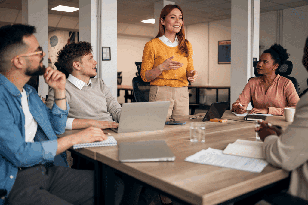Fotografia de uma mulher de blusa amarela em pé liderando uma reunião com sua equipe ao redor de uma mesa em um escritório moderno