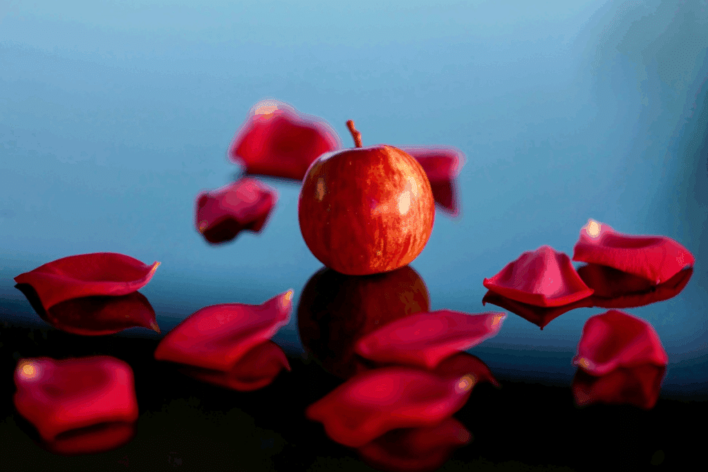 Fotografia de uma maçã vermelha cercada por pétalas de rosa vermelhas sobre uma superfície refletiva com fundo azul.