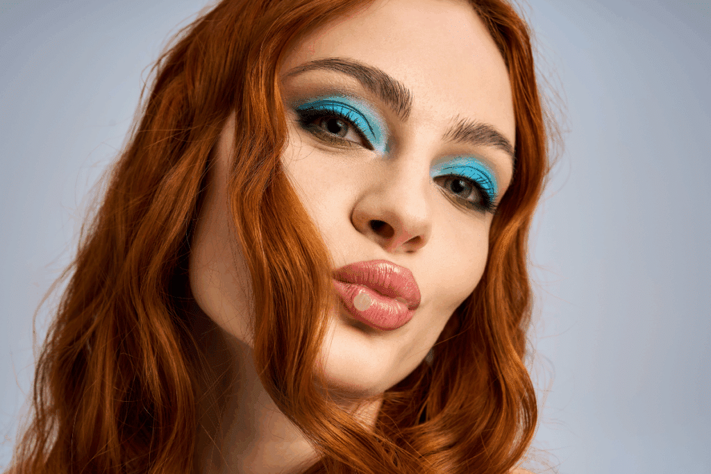 Close no rosto de uma mulher ruiva fazendo bico com os lábios. Ela usa maquiagem colorida nos olhos com sombra azul-vibrante na pálpebra e detalhes em verde rente aos cílios inferiores. O fundo é neutro.