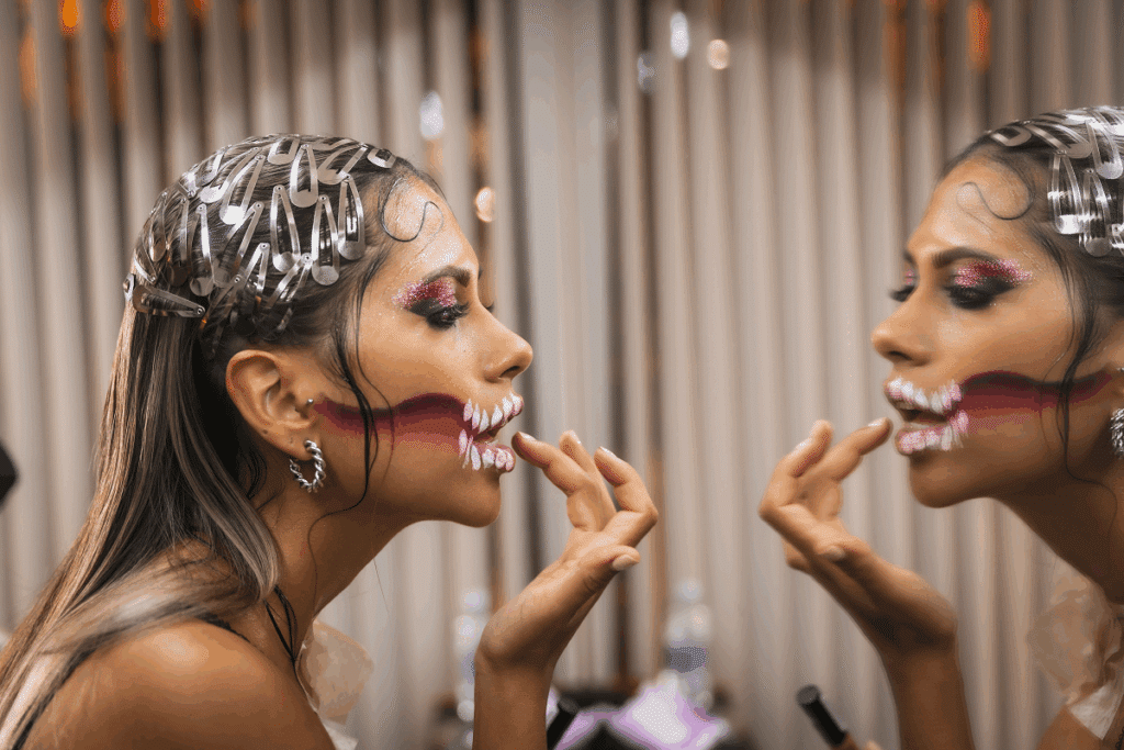 Uma mulher de perfil olha para o espelho enquanto ajusta sua maquiagem artística de Halloween, que simula uma boca rasgada com dentes pintados em tons de vermelho e branco, e traz o cabelo decorado com diversos grampos prateados
