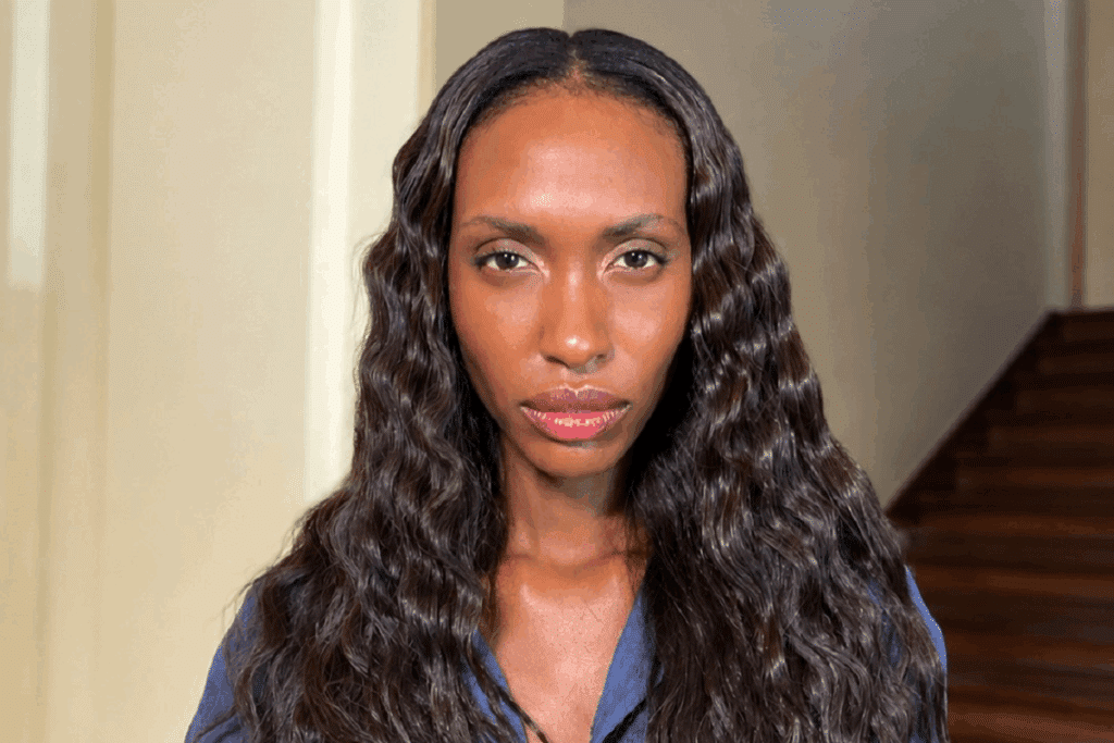 Fotografia em plano fechado de uma mulher negra de cabelos escuros, longos e ondulados, olhando diretamente para a câmera com uma expressão neutra. Ela tem lábios carnudos com acabamento brilhante e está usando uma roupa azul. Ao fundo, vê-se uma parede em tom claro e uma escada de madeira.