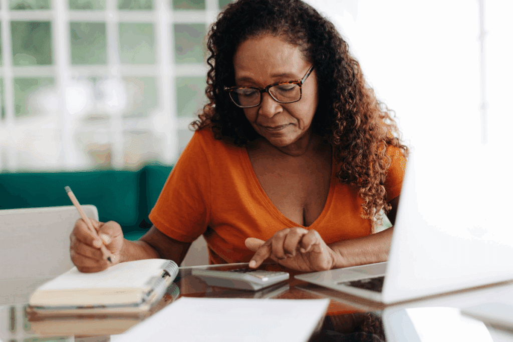 Mulher negra de óculos e cabelos cacheados sentada à mesa, utilizando uma calculadora e escrevendo em um caderno em frente a um notebook.