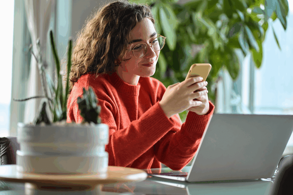 Mulher de óculos e suéter alaranjado sorri enquanto digita no celular, sentada em uma mesa de trabalho com um notebook aberto e plantas ao fundo.