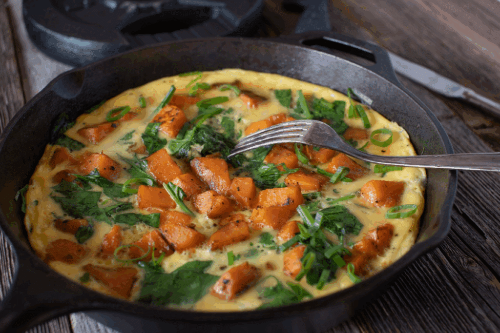 Frigideira de ferro preta sobre uma superfície de madeira, contendo uma omelete com cubos de batata-doce, folhas de espinafre e cebolinha picada, com um garfo apoiado na borda.