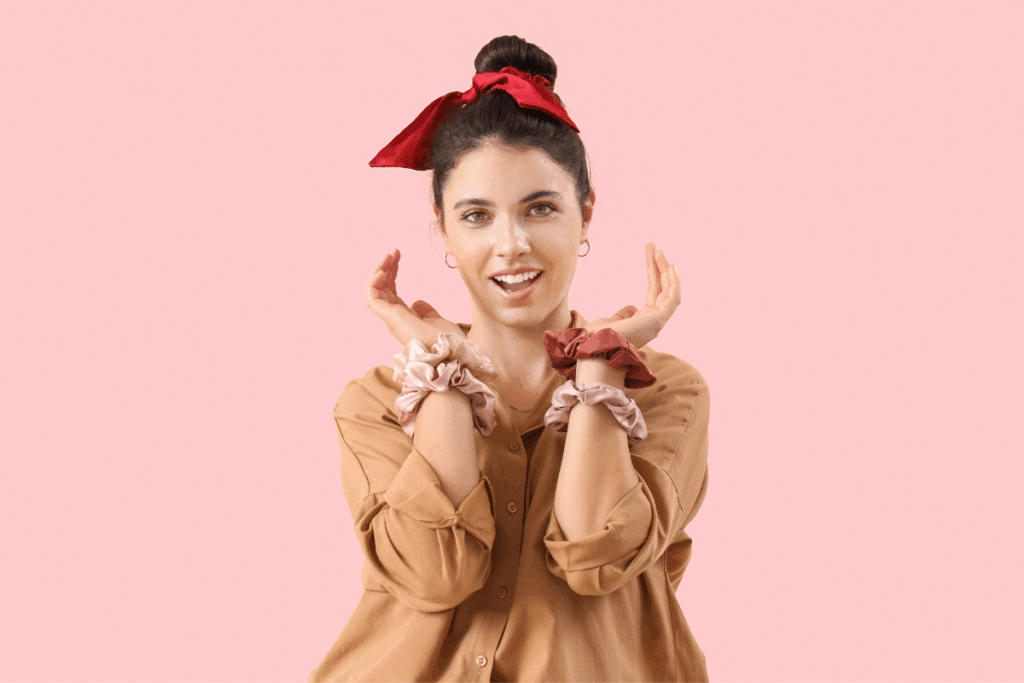 Mulher sorridente de cabelos escuros presa num coque alto com uma faixa vermelha amarada em laço. Ela veste uma camisa bege e segura os braços erguidos perto do rosto, exibindo vários elásticos de cabelo macios (scrunchies) nos pulsos, sobre um fundo rosa claro.