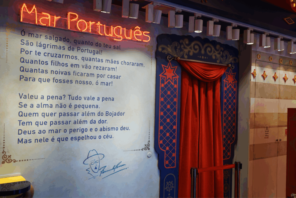 Parede clara exibindo o poema "Mar Português", de Fernando Pessoa, escrito em letras azuis. Acima do texto, destaca-se um letreiro em neon brilhante na cor laranja com o título "Mar Português". Ao lado direito da parede, há uma porta decorativa adornada com uma cortina vermelha.