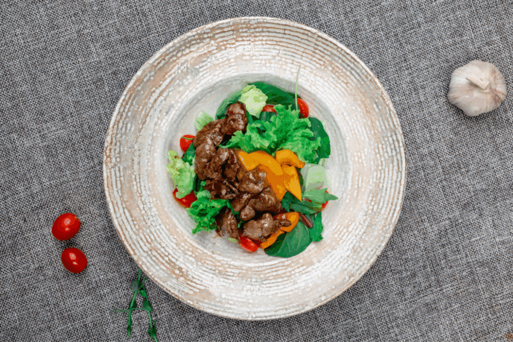 Fotografia em vista superior de uma salada servida em um prato fundo bege sobre uma superfície cinza com textura de tecido. O prato combina folhas verdes, tiras de pimentão amarelo, tomates-cereja e pedaços de fígado de frango grelhado no centro. Ao lado do prato, estão posicionados dois tomates-cereja e uma cabeça de alho.