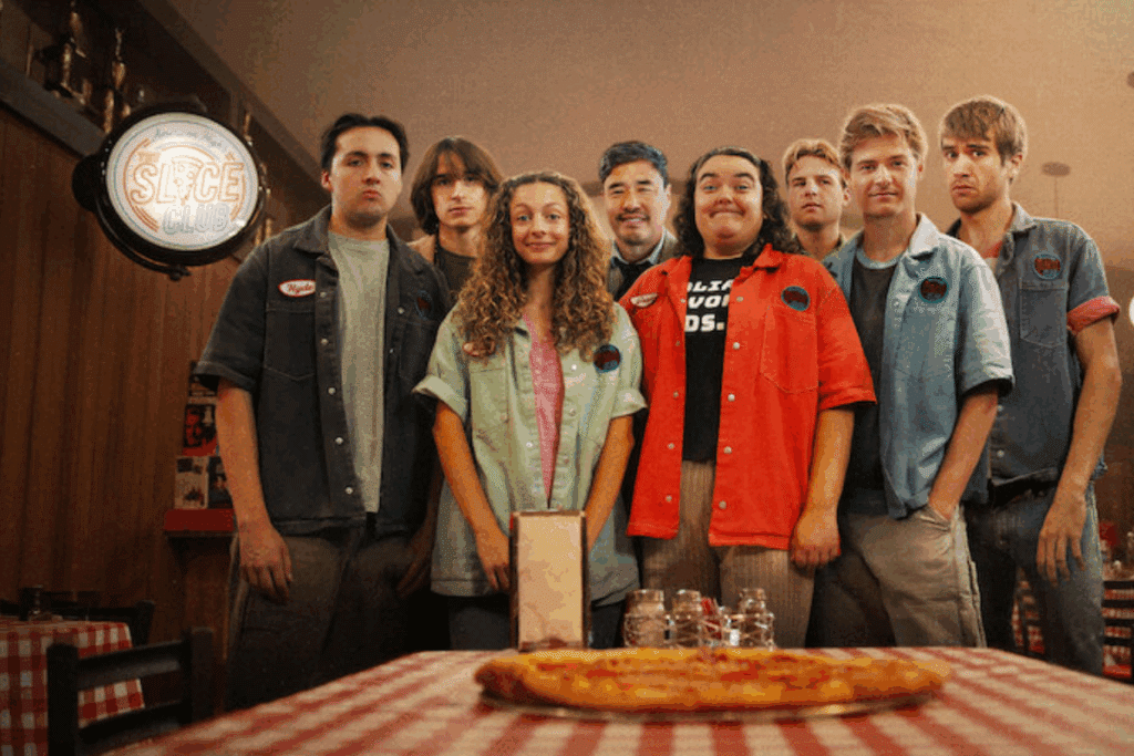 Grupo de funcionários de uniforme em uma pizzaria, posando atrás de uma mesa com uma pizza.